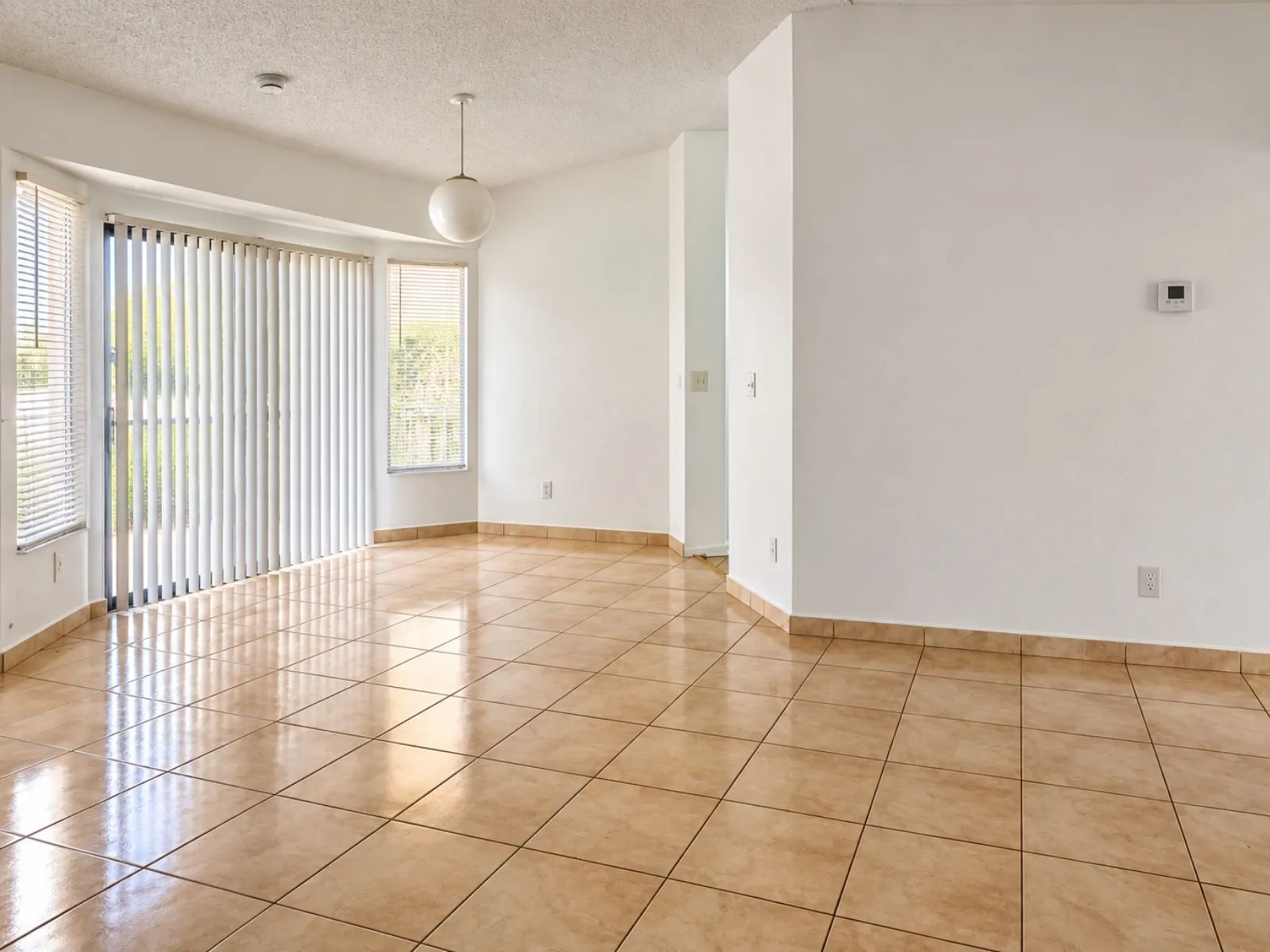 An empty room with a glossy tan tile floor, white walls and vertical blinds over a sliding window.