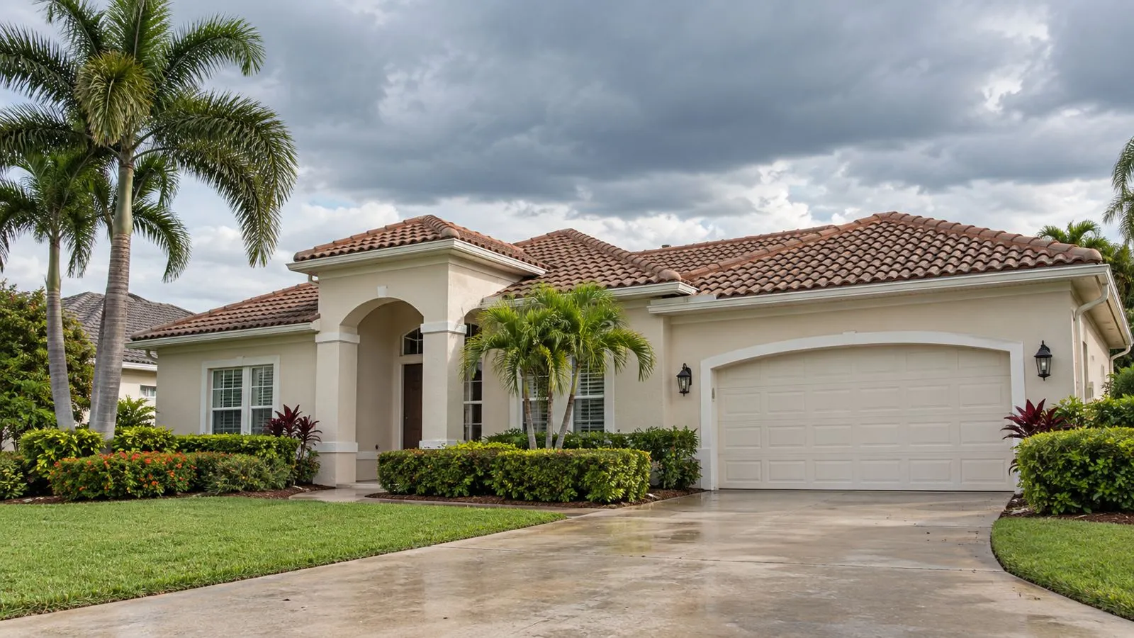 A single-story stucco house with a tile roof, palm trees and a wet driveway under a cloudy sky.