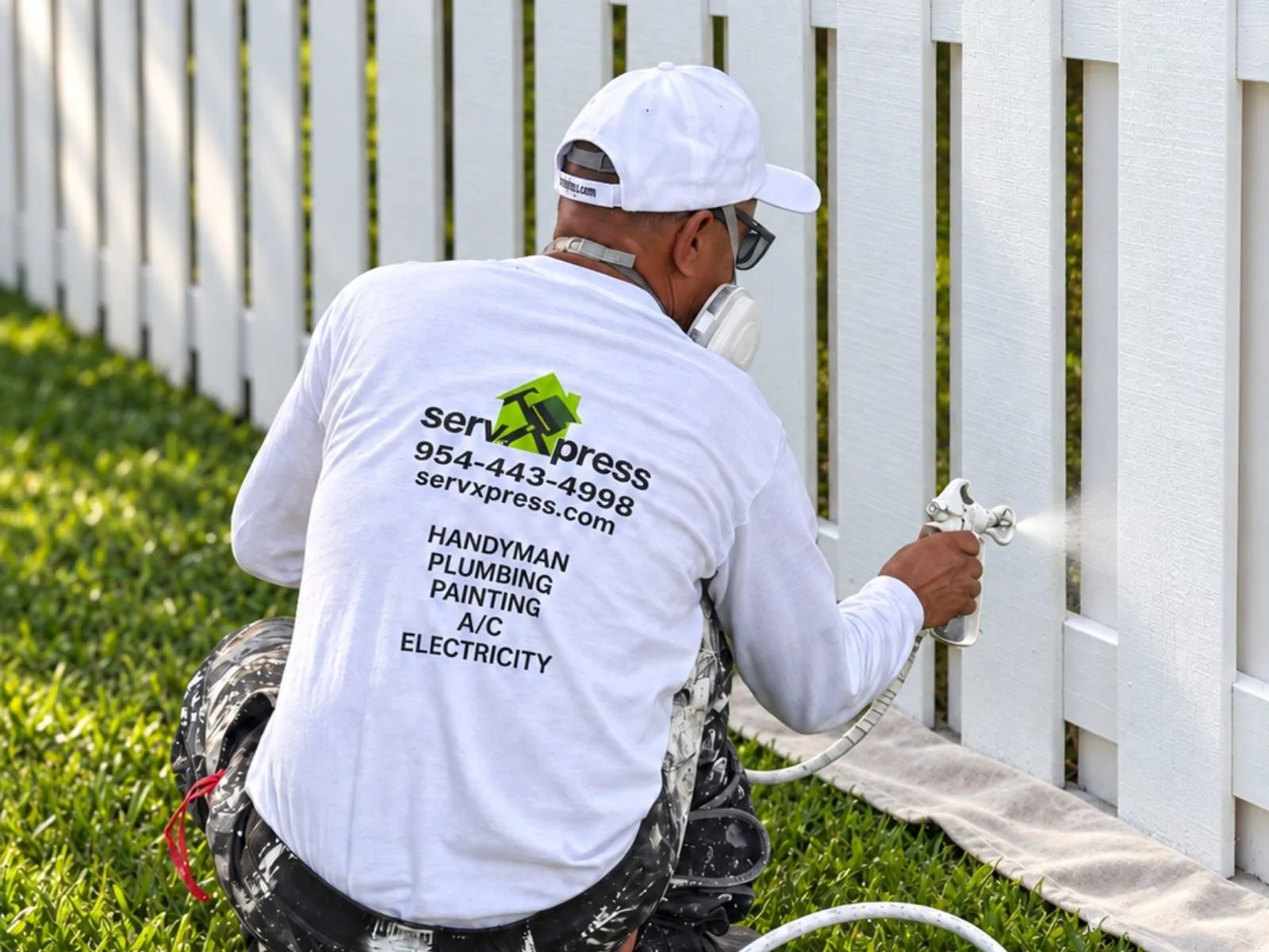 A ServXpress painter wearing a respirator and white cap spray-painting a white picket fence.