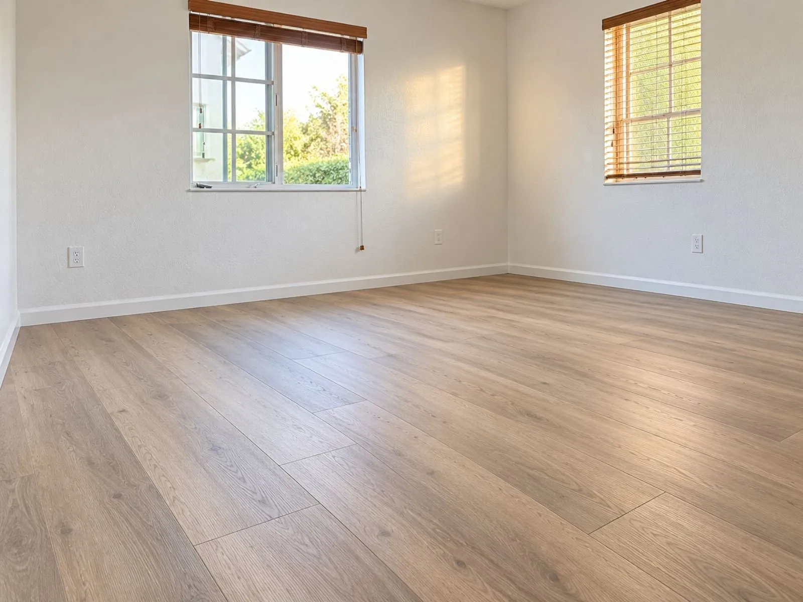 An empty room with light wood-look plank flooring, white walls and two windows with wooden blinds.